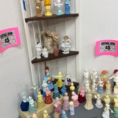 Vintage Avon figurine perfume bottles in various designs and colors displayed alongside small porcelain figurines and ceramic dishware.