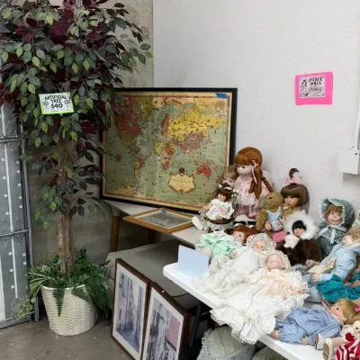 Large artificial ficus tree in a potted basket, displayed alongside framed wall art including a world map, porcelain dolls, and textiles.
