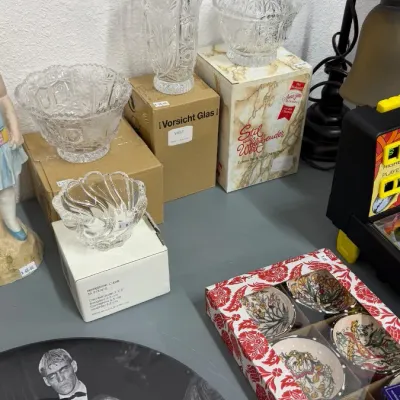 Cut crystal glassware including a covered candy dish, vases, and bowls displayed alongside boxed ceramic items and an Addams Family...