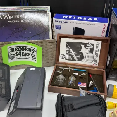 Vinyl LP records alongside an MAC Tools air-brush set in a wooden case, a Netgear N600 router, audio equipment, and a small camera bag.