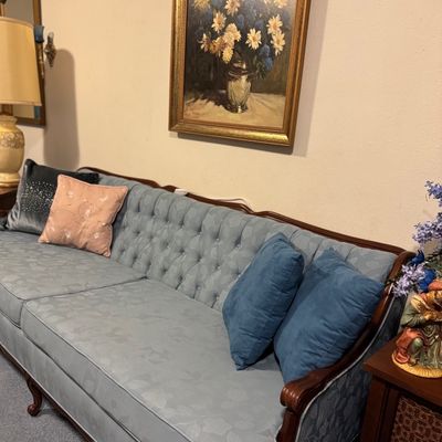 Sale Photo Thumbnail #30: Vintage French Provincial-style light blue upholstered sofa featuring a tufted backrest, wood trim, and throw pillows. Hanging above is a framed floral still life oil painting in an ornate gold frame.