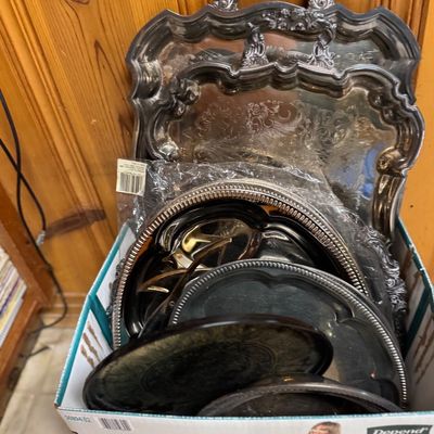 Sale Photo Thumbnail #53: Cardboard box filled with assorted vintage silver-plated serving trays and platters, including ornate footed and handled pieces with etched scrollwork.