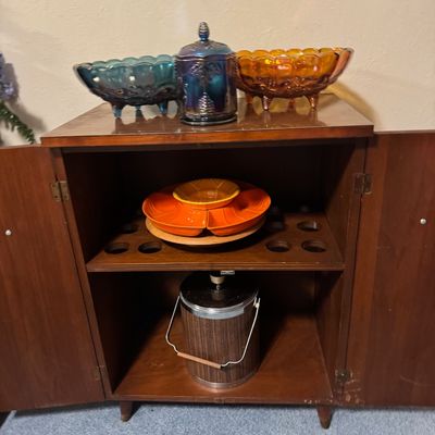 Sale Photo Thumbnail #27: Mid-century wooden dry bar cabinet filled with glassware, an orange relish set, and a faux-wood ice bucket.