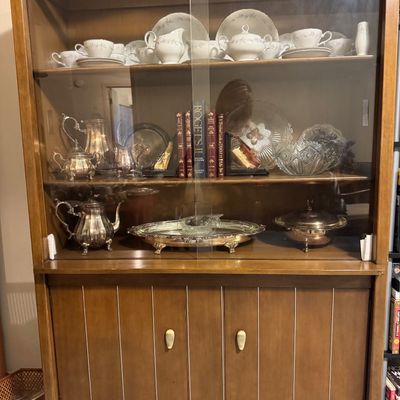 Sale Photo Thumbnail #23: Mid-century wooden hutch cabinet with glass sliding doors, featuring shelves filled with silver plate serveware, fine china, books, and glassware.