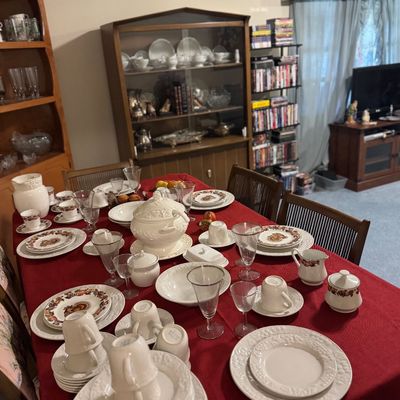 Sale Photo Thumbnail #40: Dining table set with extensive porcelain dinnerware, teacups, and stemware on a red tablecloth, alongside wooden china cabinets and a media shelf in the background.