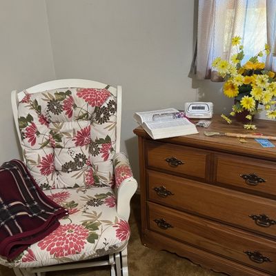 Sale Photo Thumbnail #45: White wooden glider armchair with floral cushions paired with a traditional oak three-drawer dresser and brass-base table lamp.
