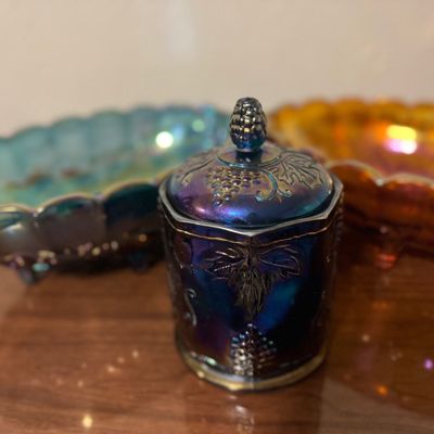 Sale Photo Thumbnail #25: Vintage iridescent purple carnival glass lidded jar with grape and cable pattern, accompanied by additional carnival glass dishes in the background.