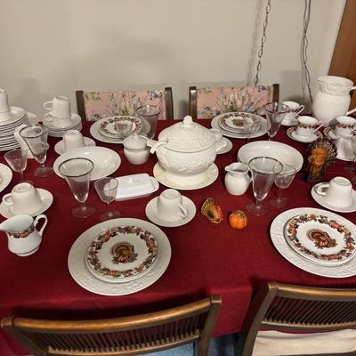 Sale Photo Thumbnail #22: Large formal dinnerware set arranged on a red tablecloth, including embossed white dishes, a tureen, glassware, and turkey-patterned Thanksgiving plates.