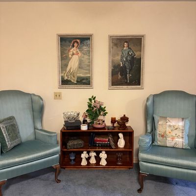 Sale Photo Thumbnail #31: Pair of light blue upholstered wingback armchairs flank a small wood bookshelf, set beneath framed prints of Thomas Gainsborough's 'The Blue Boy' and Thomas Lawrence's 'Pinkie'.