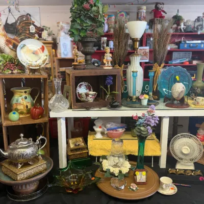 Sale Photo Thumbnail #10: An eclectic collection of home decor and tabletop items, featuring a large rooster figurine, ceramic pottery, glassware, a vintage fan, and various decorative accent pieces displayed on wooden crates and shelving.