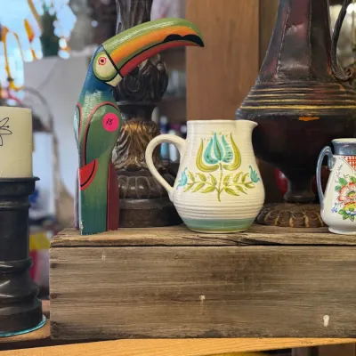 Sale Photo Thumbnail #55: Collection of decorative home accents displayed on wooden crates, including a hand-painted wooden toucan figure, floral ceramic pitchers, and a pillar candle on a dark holder.