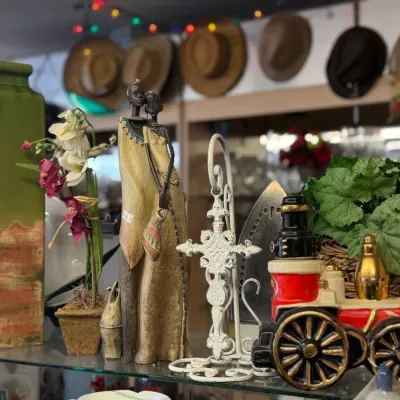 Sale Photo Thumbnail #65: An assortment of decorative home items displayed on a glass shelf, including an African tribal couple figurine, a vintage train locomotive decanter, a tall green ceramic vase, and a wrought iron cross stand.