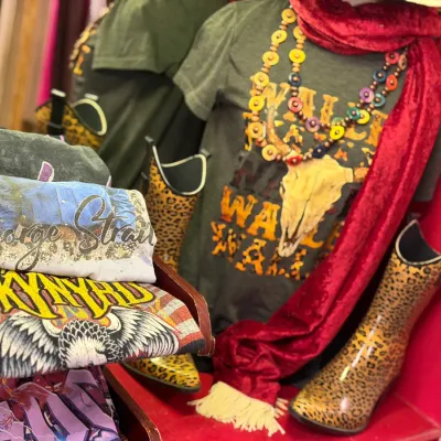 Sale Photo Thumbnail #15: Collection of Western and band apparel including folded graphic t-shirts like Lynyrd Skynyrd and George Strait, styled on a mannequin with beaded necklaces, a red scarf, and leopard print boots.