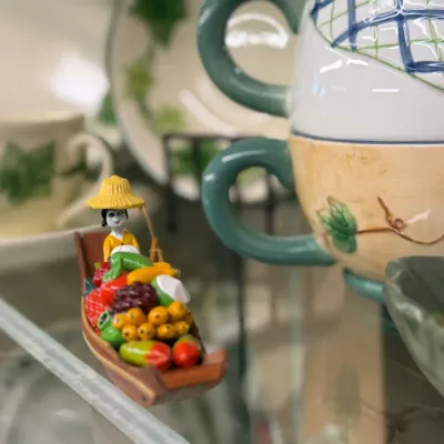 Sale Photo Thumbnail #75: Miniature hand-painted Thai floating market boat figurine filled with colorful produce, displayed alongside ceramic tea sets.