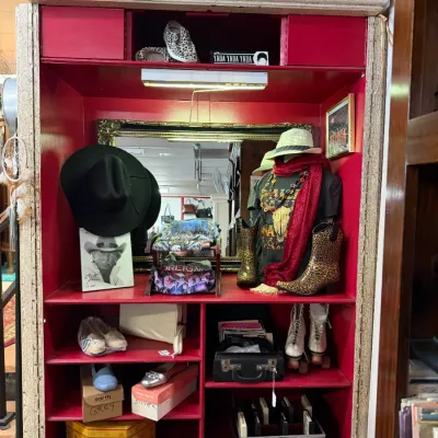 Sale Photo Thumbnail #2: Large painted wooden display hutch styled with a bright red interior, holding an assortment of vintage clothing, cowboy hats, shoes, roller skates, and decorative knick-knacks.