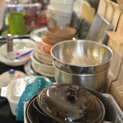 Sale Photo Thumbnail #80: Assorted cookware, mixing bowls, and kitchen supplies displayed on a table. Includes stacked metal mixing bowls, non-stick skillets with a amber glass lid, ceramic bowls, and various dishware.