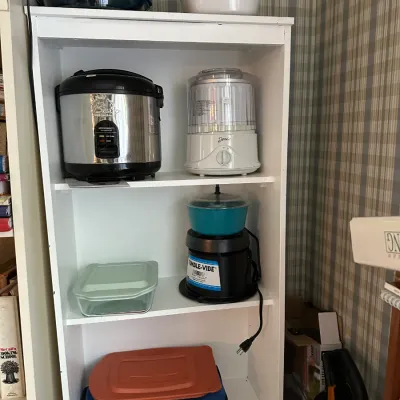 White wooden bookshelf filled with assorted kitchen appliances, CorningWare, and covered glass baking dishes, including a rice cooker and...