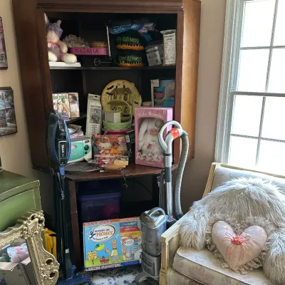 A vintage dark wood corner cabinet filled with various household items, toys, and collectibles, displayed alongside upright vacuums and...