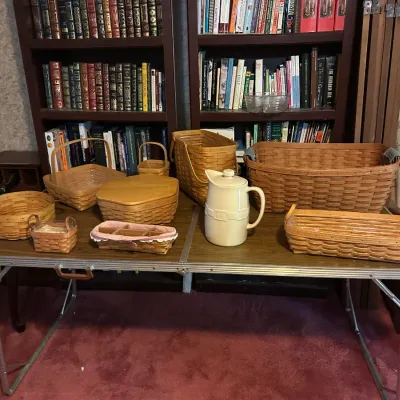Collection of Longaberger woven baskets in various shapes and sizes arranged on a folding table alongside a ceramic pitcher.