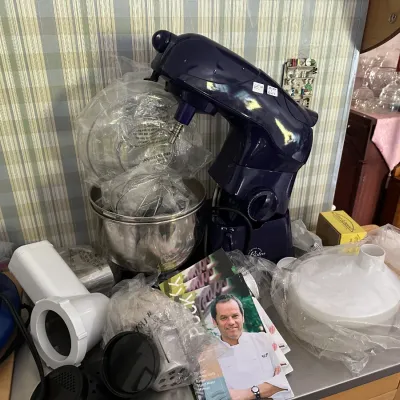 Bodum Bistro electric stand mixer in dark blue, displayed with mixing bowl, plastic-wrapped attachments, and instruction manuals.