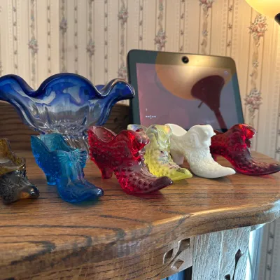 Collection of vintage glass shoe and boot figurines in various colors including amber, blue, red, yellow, and milk glass, displayed...