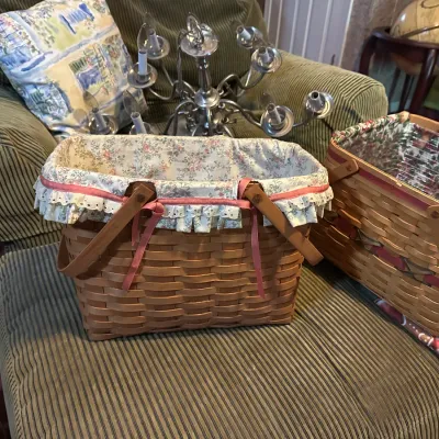 Two woven Longaberger-style picnic baskets with fabric liners and wooden swing handles, sitting on a corduroy ottoman in front of an...