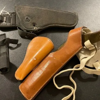 Assortment of handgun holsters including a black nylon belt holster, a tan leather shoulder holster, and a small leather sleeve, showing...