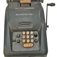 Vintage Remington Rand mechanical adding machine featuring a dark gray metal body, ten-key keypad, and hand crank lever, circa 1940s-1950s.
