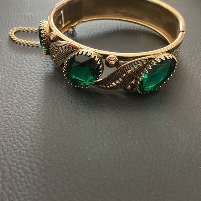 Sale Photo Thumbnail #232: Vintage gold-tone hinged bangle bracelet featuring detailed filigree and scrollwork accents set with oval green glass rhinestones. Includes a safety chain and box clasp closure. Shows light wear consistent with age, overall good vintage condition.