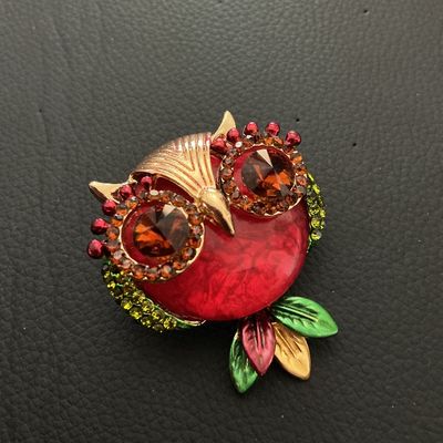 Sale Photo Thumbnail #221: Decorative owl-shaped costume brooch featuring a bold red center, amber-colored rhinestone eyes, green rhinestone accents along the sides, and multicolored leaf details. Gold-tone metal backing with standard pin closure and top loop. Measures approximatel