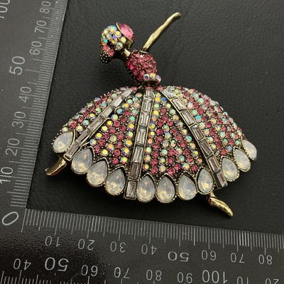 Sale Photo Thumbnail #203: Elaborate ballerina-form brooch decorated with pink, iridescent AB, and clear baguette rhinestones along with teardrop-shaped faux opalescent stones. Crafted in an antiqued gold-tone metal setting. Pin mechanism on back. Measures approximately 80mm in wid