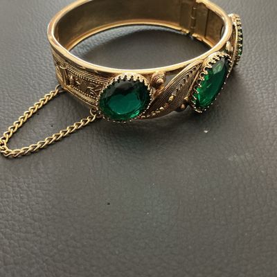 Sale Photo Thumbnail #234: Vintage gold-tone hinged bangle bracelet featuring detailed filigree and scrollwork accents set with oval green glass rhinestones. Includes a safety chain and box clasp closure. Shows light wear consistent with age, overall good vintage condition.