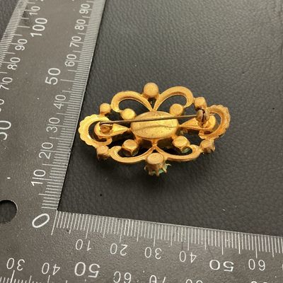 Sale Photo Thumbnail #168: Ornate vintage gold-tone openwork brooch featuring a large oval emerald-green faceted rhinestone center, accented by teardrop-cut green stones, light green cabochons, and small green rhinestone borders. Fitted with a standard pin back and rollover clasp. 