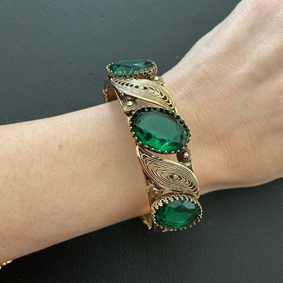 Sale Photo Thumbnail #239: Vintage gold-tone hinged bangle bracelet featuring detailed filigree and scrollwork accents set with oval green glass rhinestones. Includes a safety chain and box clasp closure. Shows light wear consistent with age, overall good vintage condition.