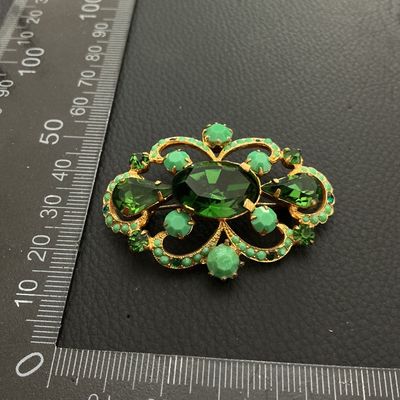 Sale Photo Thumbnail #166: Ornate vintage gold-tone openwork brooch featuring a large oval emerald-green faceted rhinestone center, accented by teardrop-cut green stones, light green cabochons, and small green rhinestone borders. Fitted with a standard pin back and rollover clasp. 