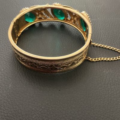 Sale Photo Thumbnail #236: Vintage gold-tone hinged bangle bracelet featuring detailed filigree and scrollwork accents set with oval green glass rhinestones. Includes a safety chain and box clasp closure. Shows light wear consistent with age, overall good vintage condition.