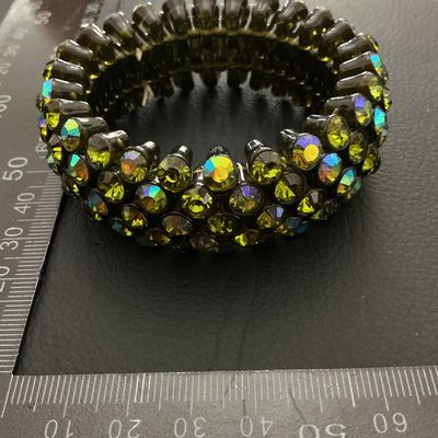 Sale Photo Thumbnail #218: A wide vintage-style stretch cuff bracelet set with multi-row olivine/olive green aurora borealis rhinestones in a dark gunmetal-tone setting. The iridescent stones display flashes of yellow, green, and blue. Measures approximately 30 mm wide with a flexi