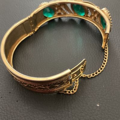Sale Photo Thumbnail #238: Vintage gold-tone hinged bangle bracelet featuring detailed filigree and scrollwork accents set with oval green glass rhinestones. Includes a safety chain and box clasp closure. Shows light wear consistent with age, overall good vintage condition.