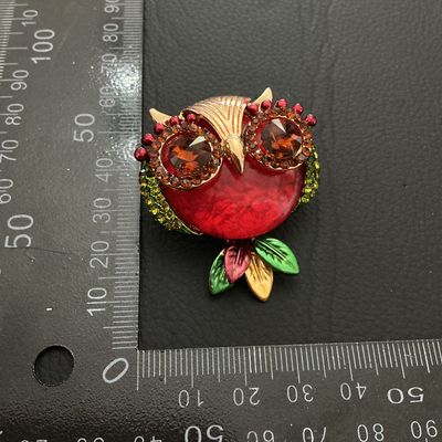 Sale Photo Thumbnail #223: Decorative owl-shaped costume brooch featuring a bold red center, amber-colored rhinestone eyes, green rhinestone accents along the sides, and multicolored leaf details. Gold-tone metal backing with standard pin closure and top loop. Measures approximatel