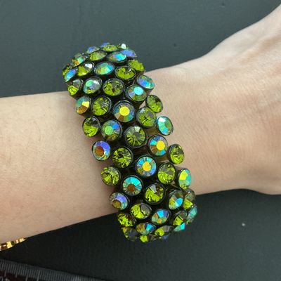 Sale Photo Thumbnail #219: A wide vintage-style stretch cuff bracelet set with multi-row olivine/olive green aurora borealis rhinestones in a dark gunmetal-tone setting. The iridescent stones display flashes of yellow, green, and blue. Measures approximately 30 mm wide with a flexi