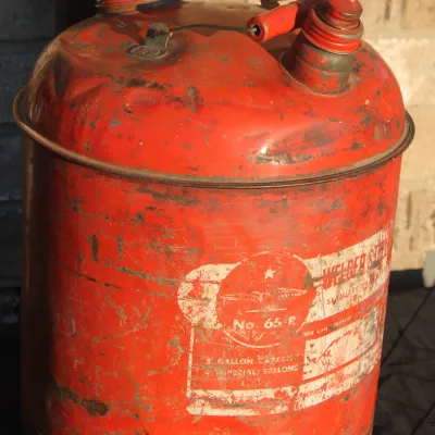 Sale Photo Thumbnail #20: Vintage red 5-gallon metal gas can featuring weathered graphics and a wooden handle grip.