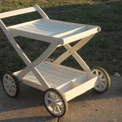 Sale Photo Thumbnail #21: White painted wooden outdoor bar cart featuring a two-tiered slatted design, built-in bottle holders, and four spoke wheels.