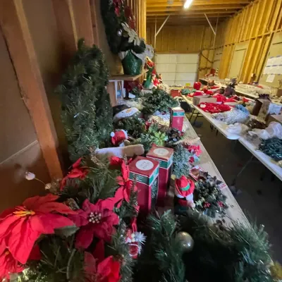 Sale Photo Thumbnail #20: Large collection of Christmas decorations, including artificial greenery, wreaths, poinsettias, and boxed ornaments, arranged on tables in a garage or workshop.
