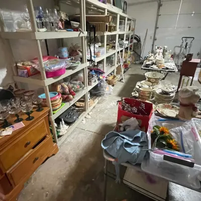 Sale Photo Thumbnail #75: Garage estate sale setup featuring plastic shelving units filled with glassware, cookware, and household goods alongside tables with stacked dishware and collectibles.