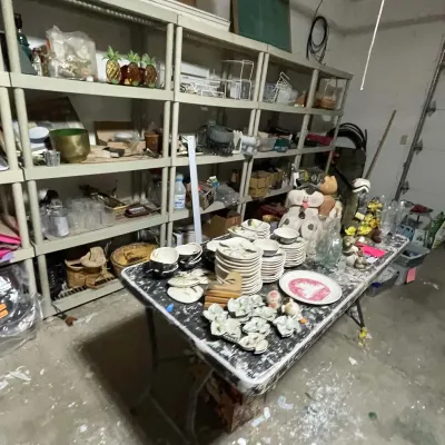 Sale Photo Thumbnail #78: Folding table and shelving units loaded with assorted household items, including dishware sets, decorative figurines, and glassware.