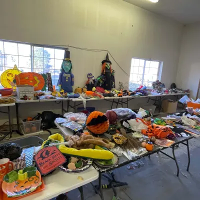 Sale Photo Thumbnail #11: Folding tables filled with Halloween decorations and party supplies, including witch figures, jack-o'-lantern items, signs, and costume accessories.