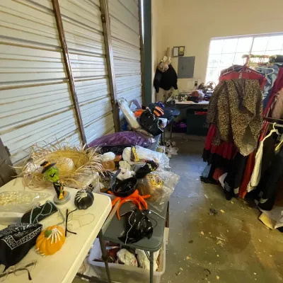 Sale Photo Thumbnail #18: Collection of Halloween costumes, decor, and accessories arranged on folding tables and clothing racks in a garage setting.