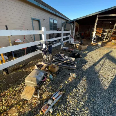 Sale Photo Thumbnail #29: Outdoor estate sale items including a portable propane grill, hydraulic floor jack, folding lawn chairs, umbrella parts, and yard tools arrayed along a white fence.