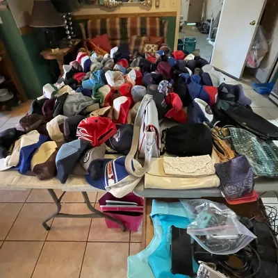 Sale Photo Thumbnail #95: A folding table covered in a large collection of vintage and modern baseball caps, trucker hats, and women's handbags.