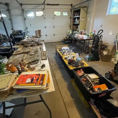 Sale Photo Thumbnail #71: Garage sale setup featuring folding tables and storage totes filled with sheet music, books, glassware, footwear, and assorted household goods.